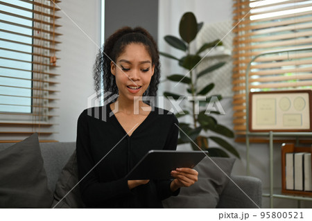 Beautiful young African woman relaxing on sofa in cozy living room and using digital tablet 95800521