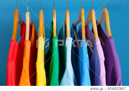 Bright clothes on wooden hangers against blue background. Rainbow colors Bright clothes on wooden hangers against blue background. Rainbow colors 95802967