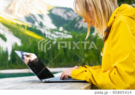 The girl is focused on working at a computer in nature with a beautiful view of the mountains and the lake. Freelancer works remotely from the office in sunny day. The girl is focused on working at a computer in nature with a beautiful view of the mountains and the lake. Freelancer works remotely from the office in sunny day. 95805414