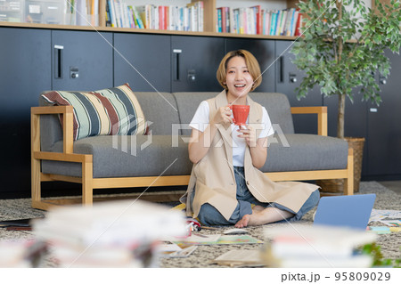 床に書類を広げて仕事しながら飲み物を飲む若い女性 95809029
