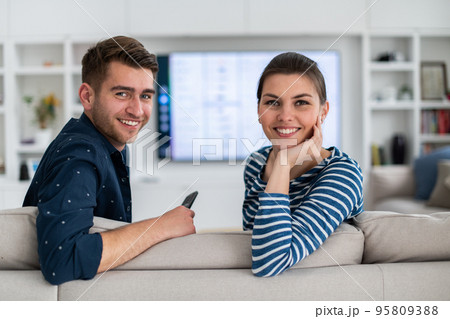 Couple Watches TV together while Sitting on a Couch in the Living Room. Girlfriend and Boyfriend embrace, cuddle, talk, smile and watch Television Streaming Services. Home with Cozy Stylish Interior. Couple Watches TV together while Sitting on a Couch in the Living Room. Girlfriend and Boyfriend embrace, cuddle, talk, smile and watch Television Streaming Services. Home with Cozy Stylish Interior. 95809388