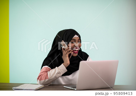 Afro woman wearing a hijab thoughtfully sitting in her home office and uses a laptop and smartphone  95809413