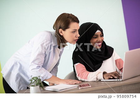 Afro woman with a hijab and a European woman using a smartphone and laptop in their home office Afro woman with a hijab and a European woman using a smartphone and laptop in their home office 95809754