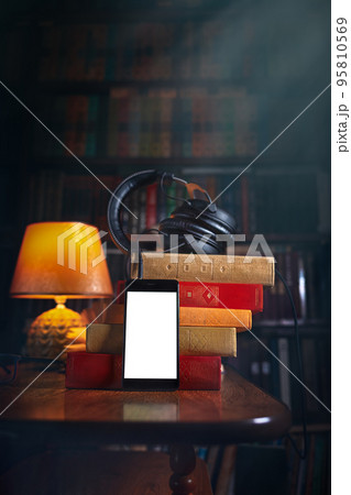 Mobile phone, headphones and a stack of books near the lamp. Concept of training and audiobooks. Library bookcase in the background. Copy space and Mock up 95810569