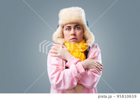 Freezing young caucasian woman wearing ushanka hat and scarf is hugging herself trying to keep warm. Gray background. The concept of flu and winter season 95810639