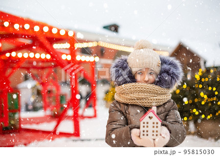 House, miniature cottage in hands of girl wearing mittens and warm clothes outdoor in snow. Family values, purchase housing, relocation, mortgage. Cozy home, Christmas, new year booking 95815030