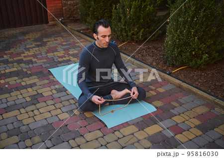 Top view of caucasian sportsman, yogi, sitting barefoot in lotus position on yoga mat, meditating with rosary. Sportsman doing breathing exercises outdoors using prayer beads. Body relaxing exercises Top view of caucasian sportsman, yogi, sitting barefoot in lotus position on yoga mat, meditating with rosary. Sportsman doing breathing exercises outdoors using prayer beads. Body relaxing exercises 95816030