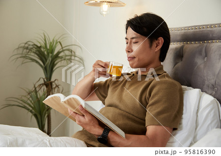 Happy calm young man drinking hebal tea and reading good book in bed Happy calm young man drinking hebal tea and reading good book in bed 95816390
