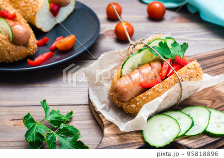 Ready-to-eat appetizing hot dog made from fried sausage, rolls and fresh vegetables, wrapped in parchment paper on a cutting board on a wooden table 95818896