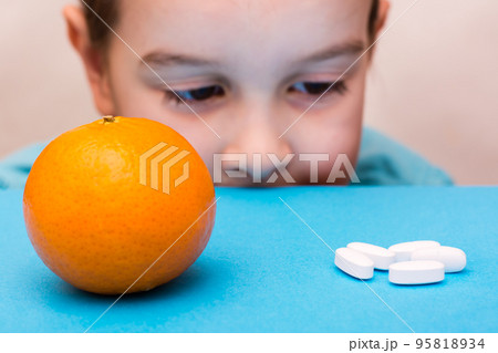 White oval pills and ripe orange lie in the face of a child on a blue background. Medicines and synthetic vitamins. The concept of choosing medicines and natural fruits. Treatment for children. 95818934