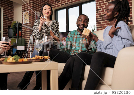 African american couple exchanging gift box during young adult home gathering, drinking alcohol. Multicultural friends group giving birthday presents to woman, enjoying. 95820180