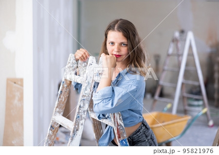Attractive young woman in denim shirt and shorts posing next to stepladder in refurbished room 95821976