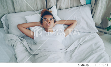 Sad race lady is lying in bed and sighing looking at ceiling then closing her eyes and falling asleep with smile. Comfortable bed, soft pillows and sheets are visible. Sad race lady is lying in bed and sighing looking at ceiling then closing her eyes and falling asleep with smile. Comfortable bed, soft pillows and sheets are visible. 95828021