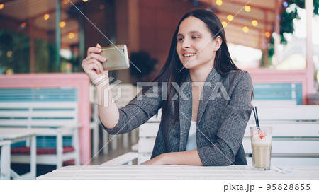 Elegant young woman is making online call using smartphone relaxing in open-air cafe at table with milkshake and talking looking at screen. Urban lifestyle, youth and technology concept. 95828855