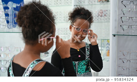 Happy African child girl testing new glasses at optical store. Eyesight and vision concept Happy African child girl testing new glasses at optical store. Eyesight and vision concept 95834324