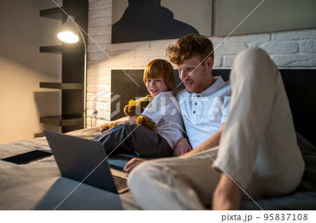 Father and son lying on bed and watching something on internet before going to sleep Father and son lying on bed and watching something on internet before going to sleep 95837108