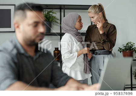 Young blond businesswoman discussing male colleague with black woman in hijab 95837696
