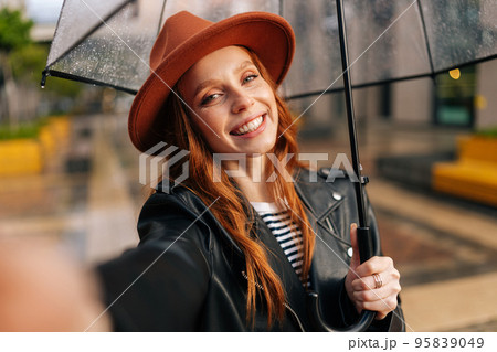 Smiling redhead young woman wearing elegant hat under transparent umbrella making selfie photo looking at camera, holding mobile phone on hand, standing in rain on European city street 95839049