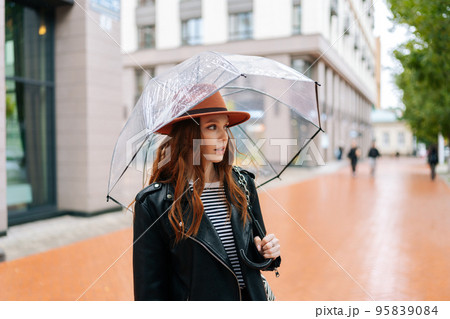 Portrait of attractive redhead young woman wearing fashion hat standing with transparent umbrella on city street near center in rainy weather rain, thinking looking away, on background of building. 95839084
