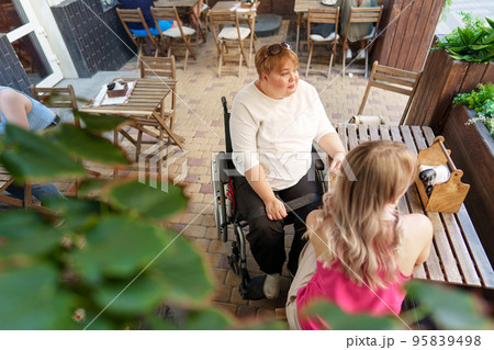 Mother with disability in wheelchair talking to her daughter while sitting at the table in cafe Mother with disability in wheelchair talking to her daughter while sitting at the table in cafe 95839498