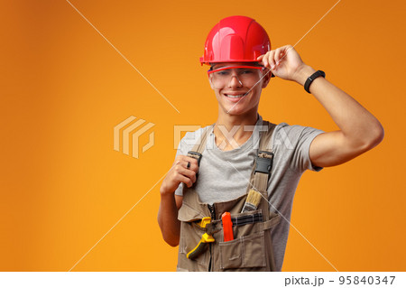 Young construction worker in helmet and uniform posing on yellow background in studio 95840347