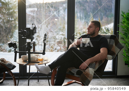 Videographer man resting on armchair, drinking tea near his camera mounted on gimbal stabilizer equipment. Videographer man resting on armchair, drinking tea near his camera mounted on gimbal stabilizer equipment. 95840549