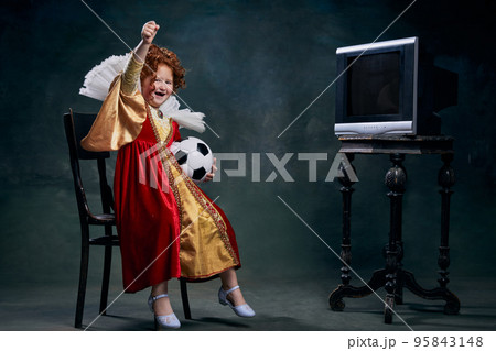 Portrait of little red-headed girl, child in costume of royal person watching football match isolated over dark green background 95843148