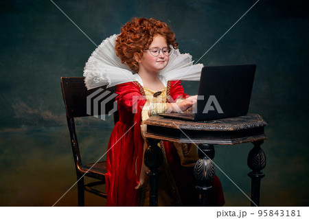 Portrait of little red-headed girl, child in costume of royal person typing on laptop isolated over dark green background Portrait of little red-headed girl, child in costume of royal person typing on laptop isolated over dark green background 95843181