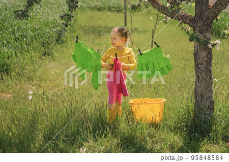 A child hangs Bright colored wet clothes on a clothesline in the garden under a tree on a sunny day. A little girl helps with household chores 95848584