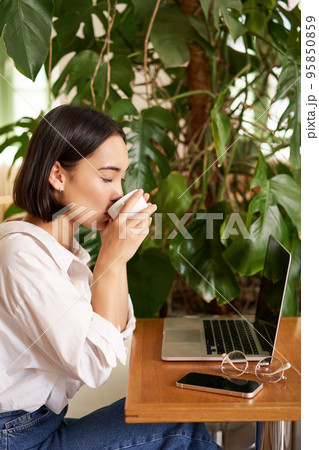 Young female entrepreneur, girl sits with laptop in cafe, works on remore from restaurant, drinks coffee and smiles 95850859