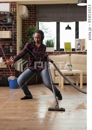 Positive housewife enjoying spring cleaning at home, singing while vacuuming in living room. Listening to music with wireless headphones relaxed looking cheerful, taking care of finishing household Positive housewife enjoying spring cleaning at home, singing while vacuuming in living room. Listening to music with wireless headphones relaxed looking cheerful, taking care of finishing household 95853023
