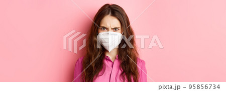 Covid-19, quarantine and social distancing concept. Close up portrait of angry female in respirator frowning, looking upset and judgemental, pink background 95856734
