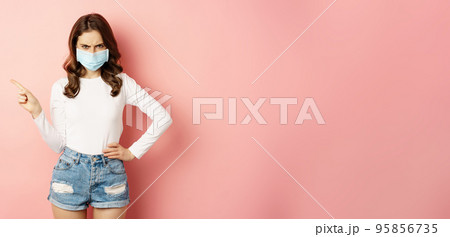 Angry young woman in medical face mask, frowning frustrated, pointing finger left, showing promo copy space, standing over pink background 95856735