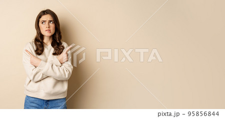 Complicated and indecisive woman pointing sideways, cant make choice, struggle with decision, choosing between two options, standing over beige background 95856844