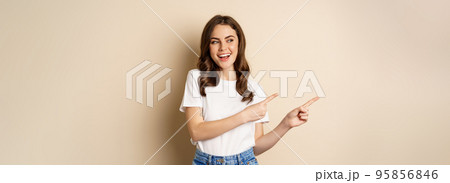 Stylish young caucasian woman smiling, pointing fingers left, showing advertisement, promo offer, standing against beige background Stylish young caucasian woman smiling, pointing fingers left, showing advertisement, promo offer, standing against beige background 95856846