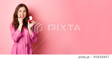 Beautiful girl dream of her wedding day, looking thoughtful up, showing engagement ring in red box and smiling, standing over pink background Beautiful girl dream of her wedding day, looking thoughtful up, showing engagement ring in red box and smiling, standing over pink background 95856847