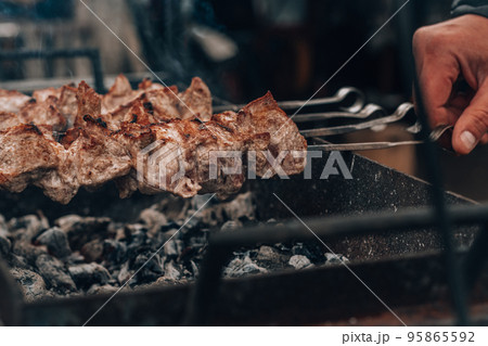 Male hand turns raw fried meat on skewers, cooked on charcoal grill. Cooking barbecue outdoors 95865592