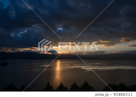 雲間から顔を出し湖面と雲を照らす夕日 95866311