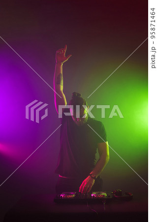 DJ at work mixing sound on her decks at a party or night club with colourful smoke light background DJ at work mixing sound on her decks at a party or night club with colourful smoke light background 95871464