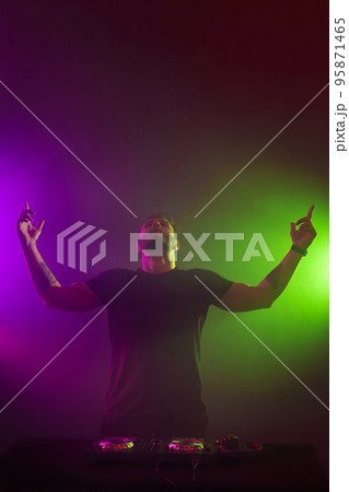 DJ at work mixing sound on her decks at a party or night club with colourful smoke light background DJ at work mixing sound on her decks at a party or night club with colourful smoke light background 95871465
