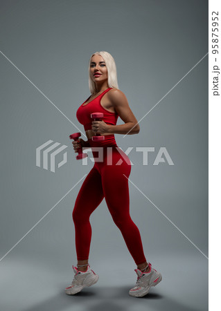 Sexy fit blonde model in red sportswear posing with small pink dumbbells 95875952