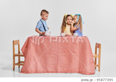 Portrait of three children, boy and girls sitting at the table over grey background. Girls whispering secrets 95877395