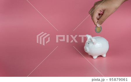 A human hand is dropping a coin into a white piggy bank. on a pink background. Savings and investment concepts. 95879061