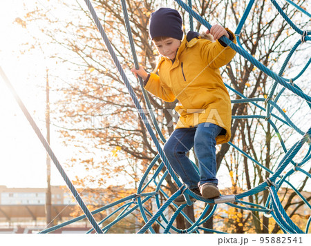 Little boy plays on children playground. Kid climbs on rope structure on sports field. Leisure activity at sunny autumn day. Fall season at town. Little boy plays on children playground. Kid climbs on rope structure on sports field. Leisure activity at sunny autumn day. Fall season at town. 95882541