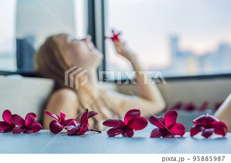 Young woman sits in a bath with foam and frangipani flowers against the background of a panoramic window overlooking the skyscrapers and a big evening city 95885987