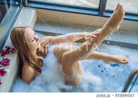 Young woman sits in a bath with foam and frangipani flowers against the background of a panoramic window overlooking the skyscrapers and a big evening city Young woman sits in a bath with foam and frangipani flowers against the background of a panoramic window overlooking the skyscrapers and a big evening city 95886007