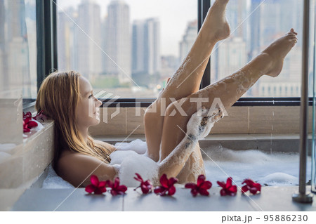 Young woman sits in a bath with foam and frangipani flowers against the background of a panoramic window overlooking the skyscrapers and a big evening city Young woman sits in a bath with foam and frangipani flowers against the background of a panoramic window overlooking the skyscrapers and a big evening city 95886230