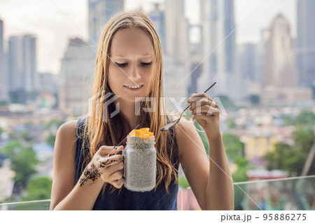 Young woman with mehendi from henna on hand eating chia pudding on her balcony overlooking the big city 95886275