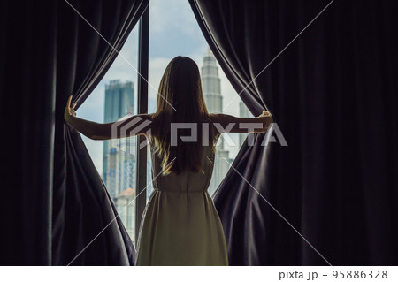 Young woman opens the window curtains and looks at the skyscrapers in the big city 95886328