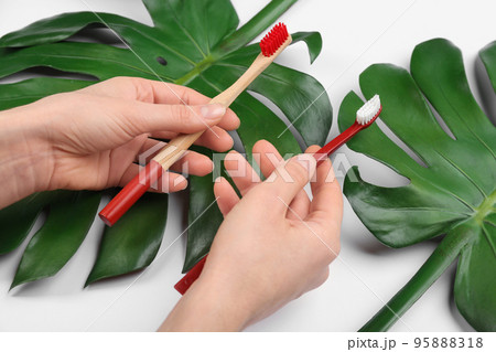 Woman holding natural bamboo and plastic toothbrushes above tropical leaves on white background, closeup Woman holding natural bamboo and plastic toothbrushes above tropical leaves on white background, closeup 95888318
